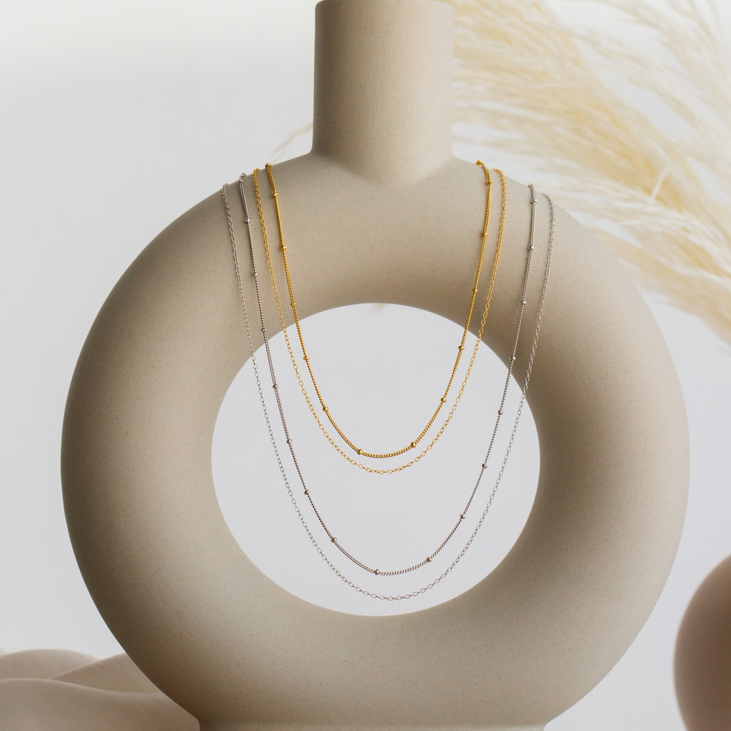 The Beaded Duo Chain Necklace, a minimalist piece, drapes elegantly over a contemporary circular beige ceramic vase.