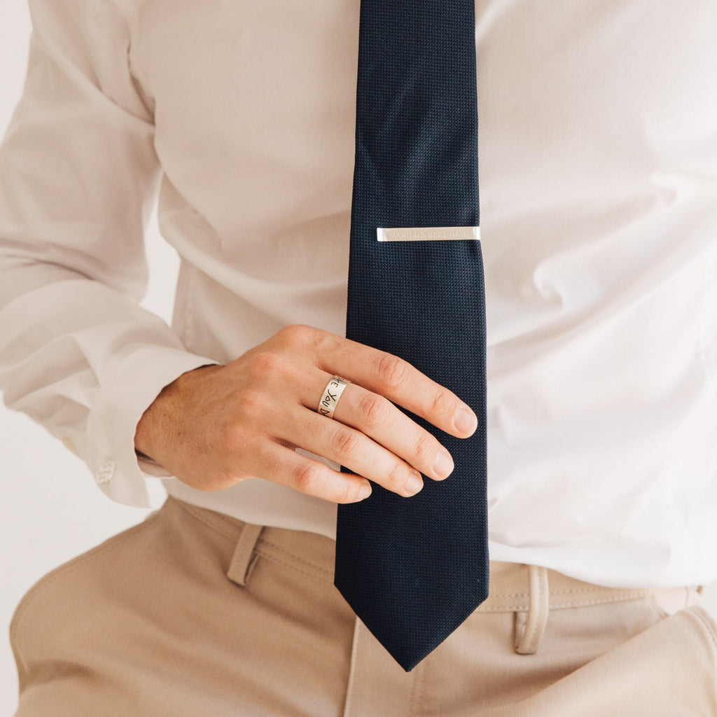 A person wearing a white shirt, beige pants, black tie, Thick Handwriting Band, and minimalist jewelry such as a silver ring and tie clip.
