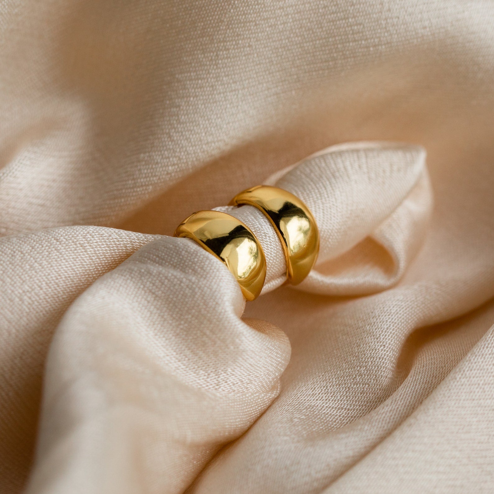 Two Olivia Hoops in 18K gold rest on soft beige satin with gentle folds, bringing a touch of contemporary style to this elegant scene.