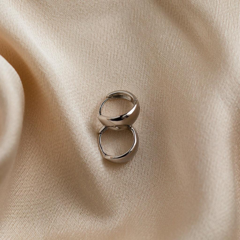 Two Olivia Hoops in Sterling Silver rest on smooth, beige satin fabric with soft folds, creating a chic and elegant scene.