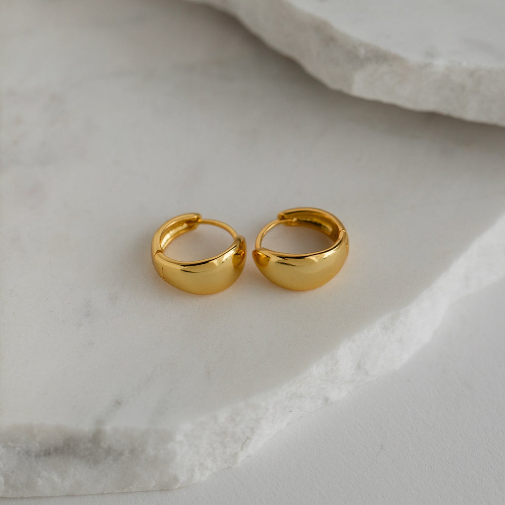 The Olivia Hoops in 18K Gold rest on a white marble surface, making them the perfect choice to elevate any contemporary look.