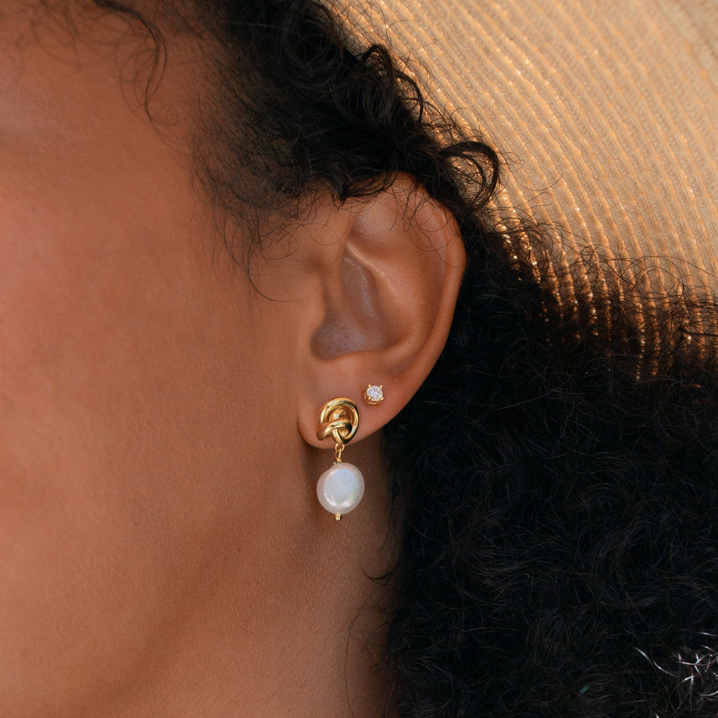 Close-up of a woman's ear wearing Knot Pearl Drop Earrings—featuring gold knots with pearl drops—and a small stud above, ideal for those who appreciate minimalist style.