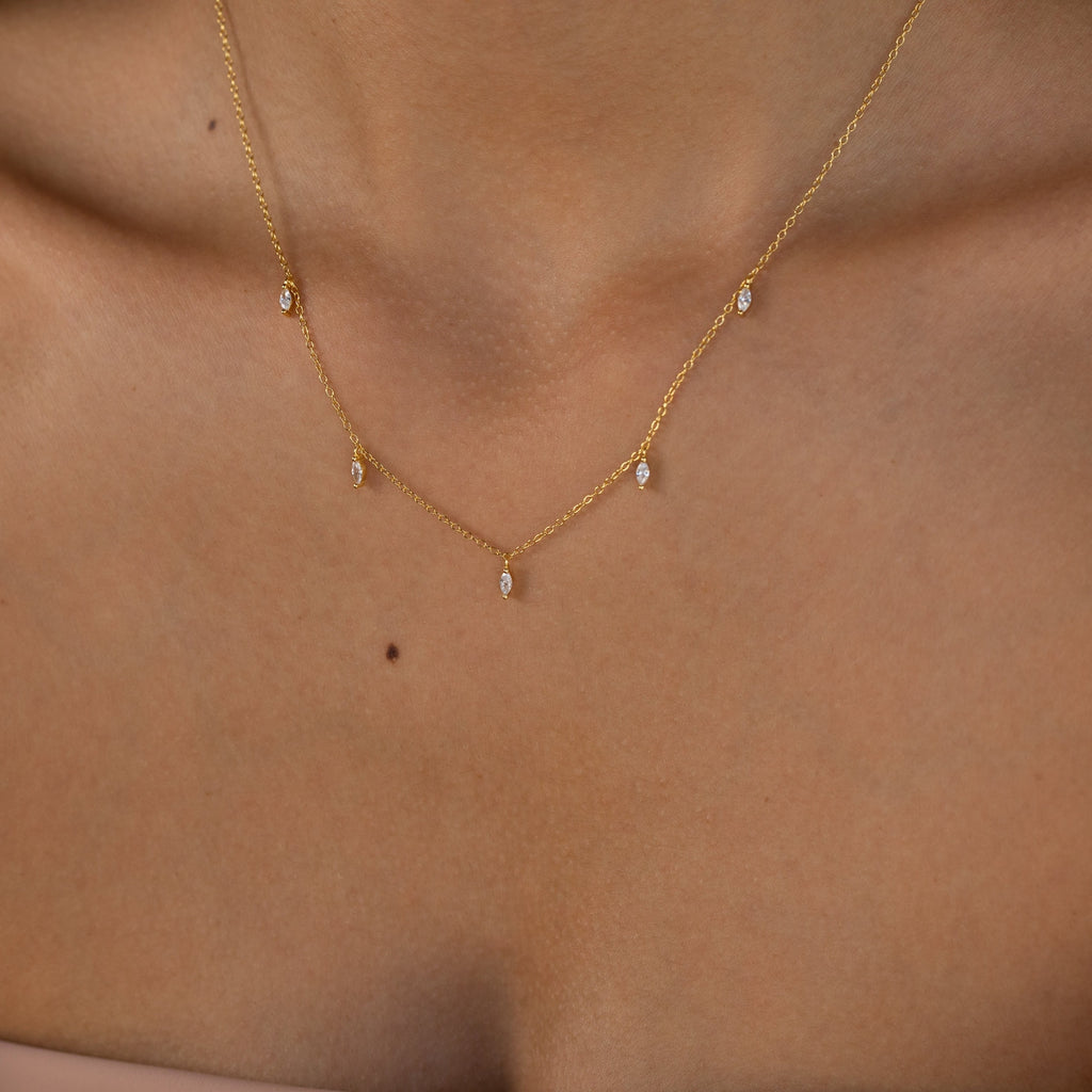 The Marquise Station Necklace gently drapes along a woman's neck and collarbone, featuring a dainty gold chain accented with marquise diamonds and dangling teardrop charms for an elegant look.