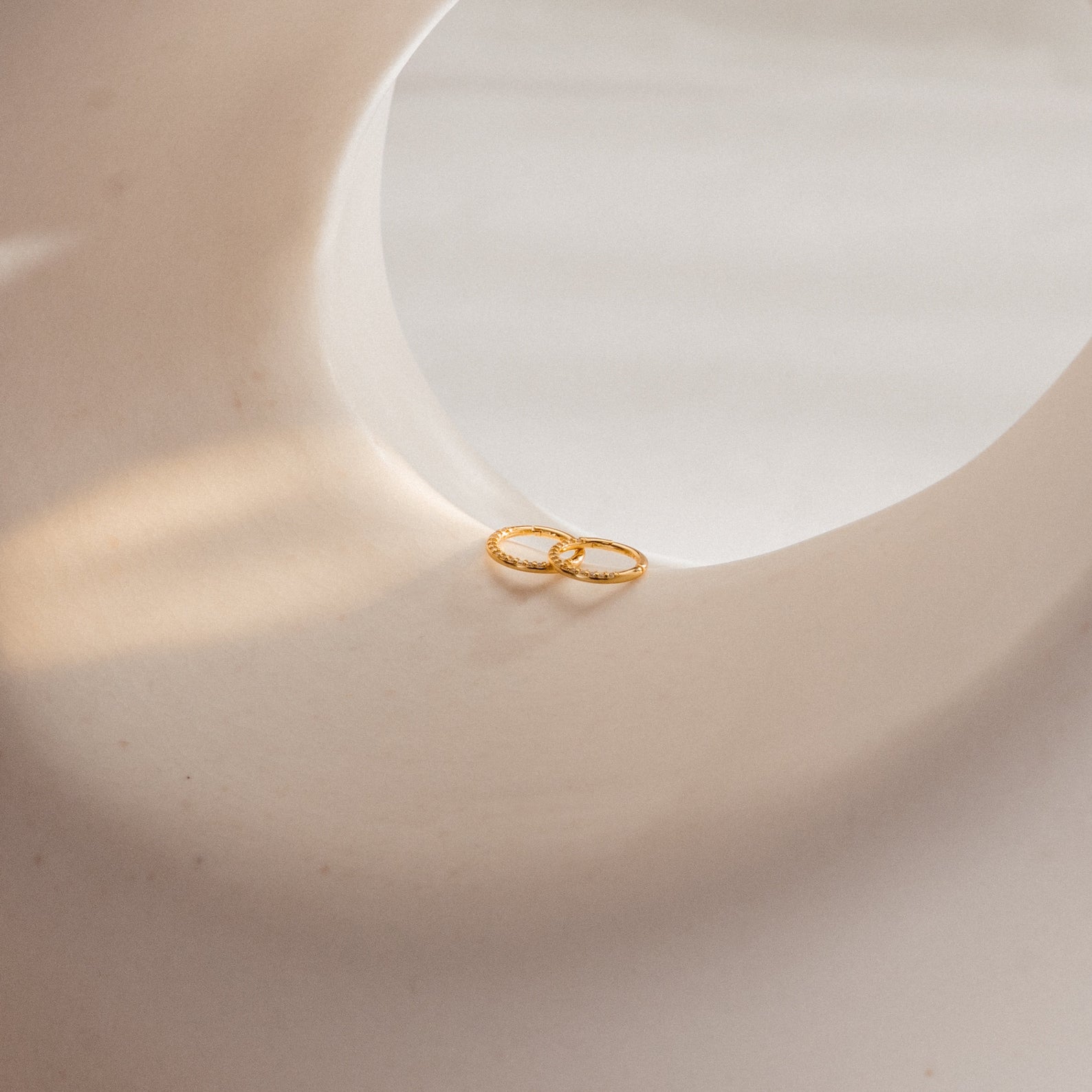 Pave Cartilage Mini Huggies in gold rest on a smooth, curved white surface under soft, natural lighting.