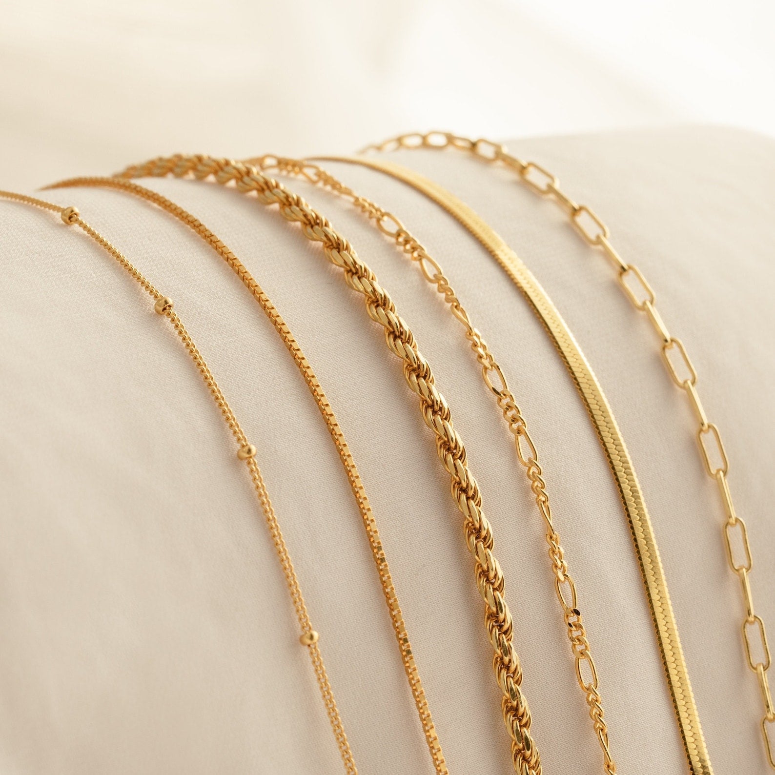 Six gold chain necklaces in various link styles, displayed on a white cushioned surface, pair stylishly with Must-Have Bracelet Chains for a chic, coordinated look.
