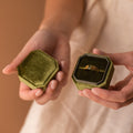 Hands holding an open green Velvet Ring Box with a gold ring inside, ideal as a jewelry gift box, shown against a neutral background.