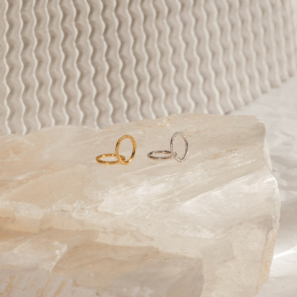 Two delicate rings, one gold and one silver, rest on textured stone with a wavy background—styled with our Pave Cartilage Mini Huggies in Sterling Silver for a refined, modern look.
