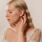 A woman with long blonde hair in a sheer white top touches her ear, highlighting the delicate Pave Cartilage Mini Huggies in Sterling Silver as she looks down with her eyes closed.