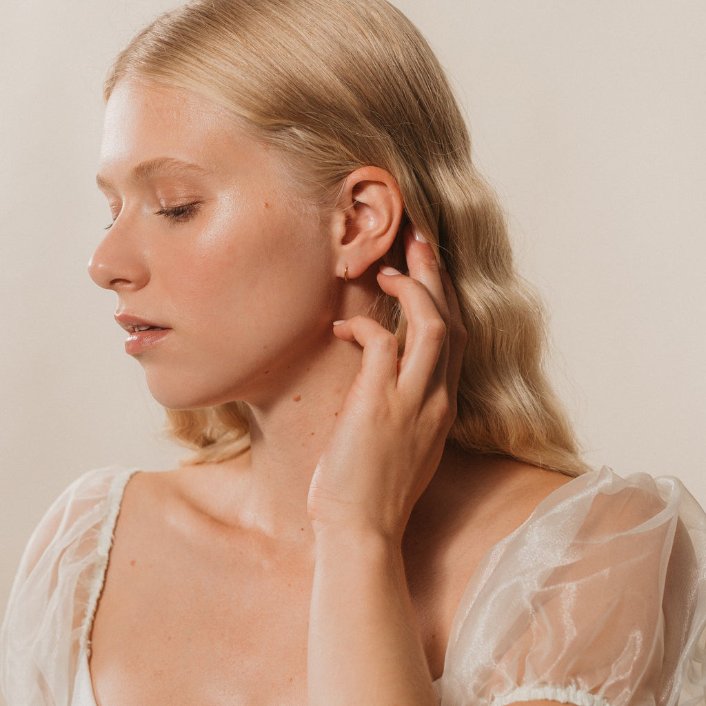 A woman with blonde wavy hair in a sheer white top touches her ear, showing off the Pave Cartilage Mini Huggies, as she faces left with her eyes closed.
