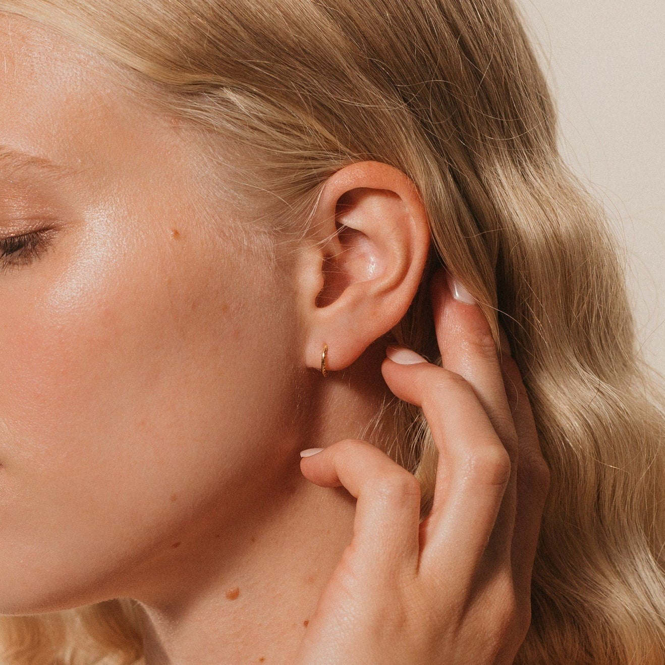 Close-up of a blonde woman touching her ear, wearing elegant Pave Cartilage Mini Huggies and a small gold hoop earring.