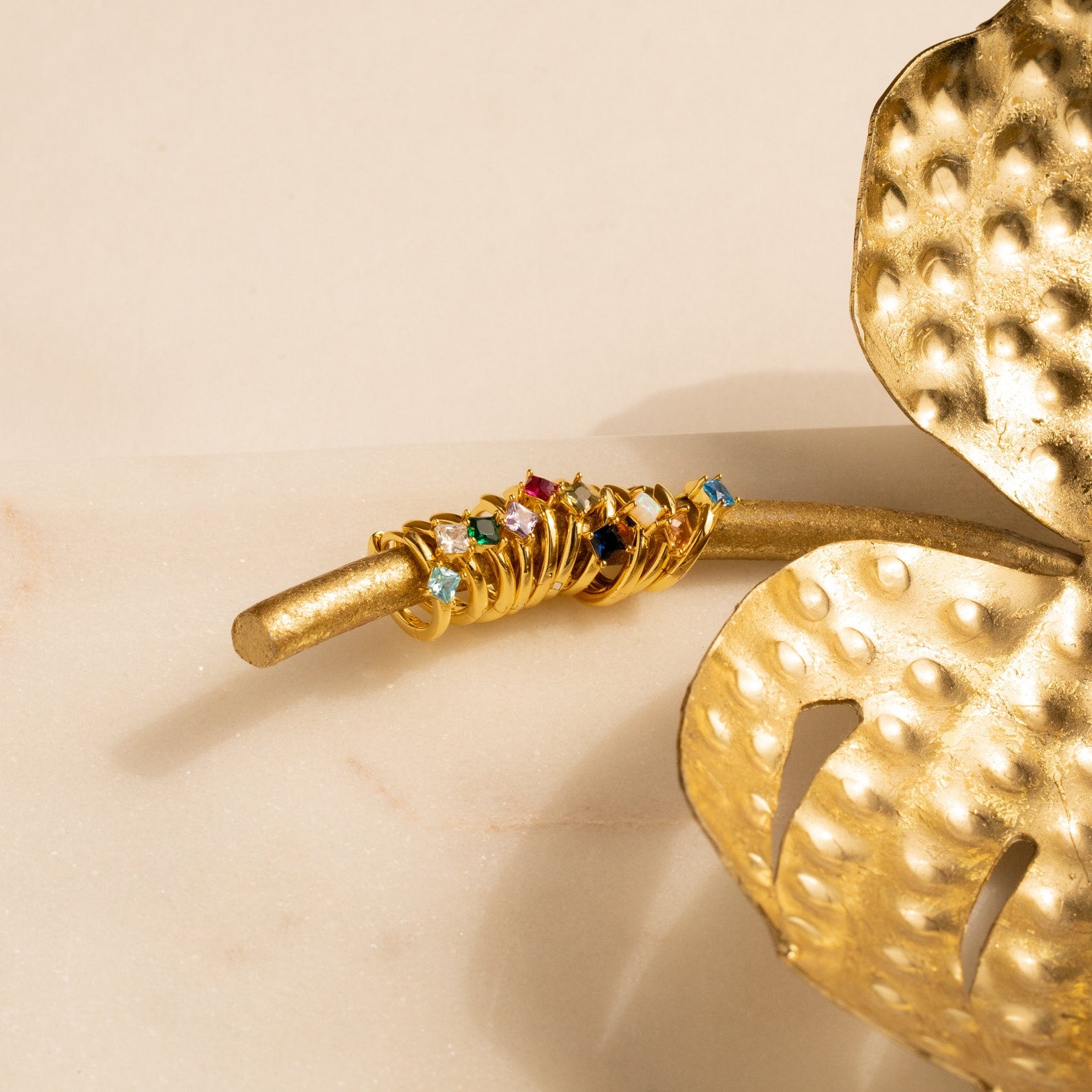 Gold rings with colorful gemstones stacked on a golden branch beside a decorative golden leaf, perfect for pairing with personalized jewelry or Athena Birthstone Huggies.