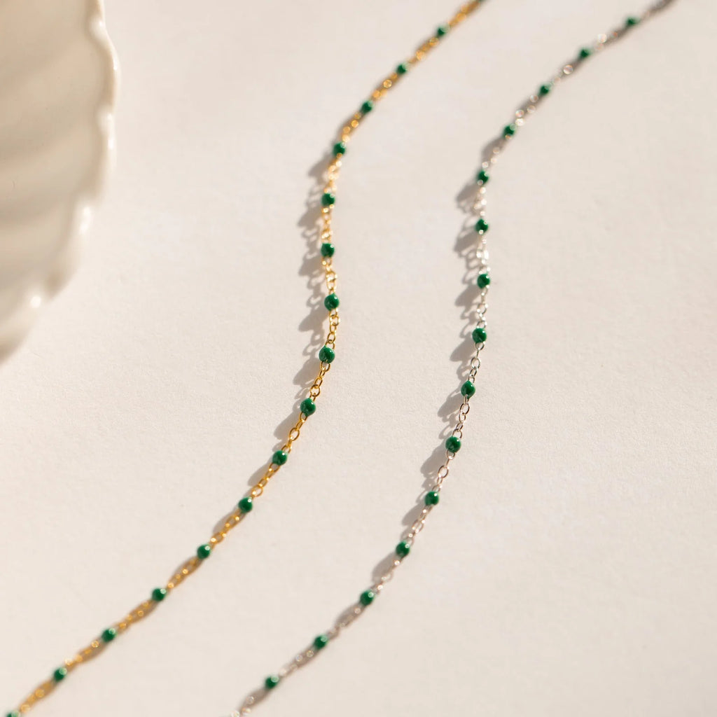 The Evergreen Necklace Set features two delicate chains with green stones—one gold, one silver—elegantly displayed on a neutral background.