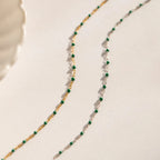 The Evergreen Necklace Set features two delicate chains with green stones—one gold, one silver—elegantly displayed on a neutral background.
