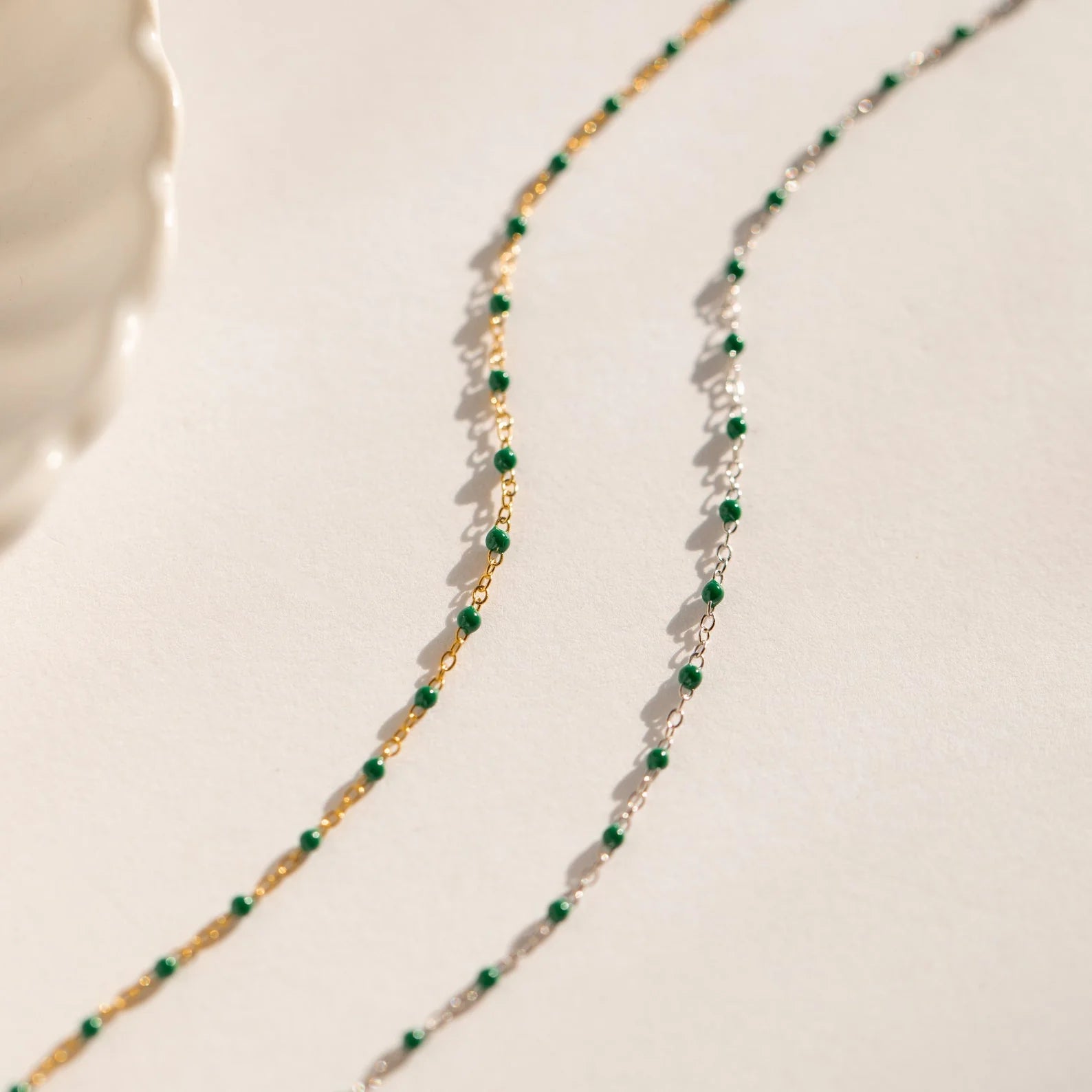 The Evergreen Necklace Set features two delicate chains with green stones—one gold, one silver—elegantly displayed on a neutral background.