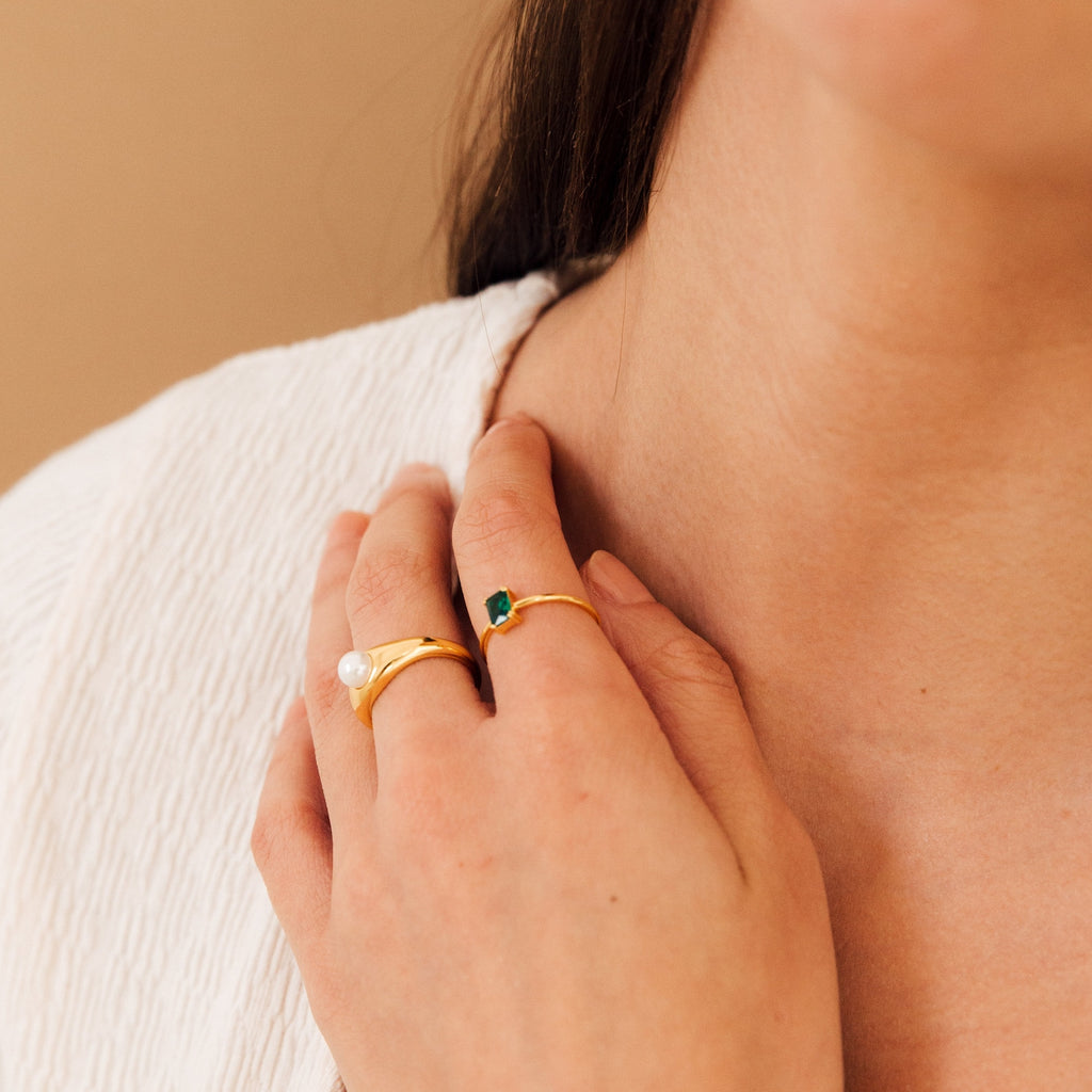 A woman touches her collarbone, showcasing vintage-style jewelry: two rings—a gold ring with a pearl and the Harley Emerald Ring in silver—evoking Art Deco elegance.