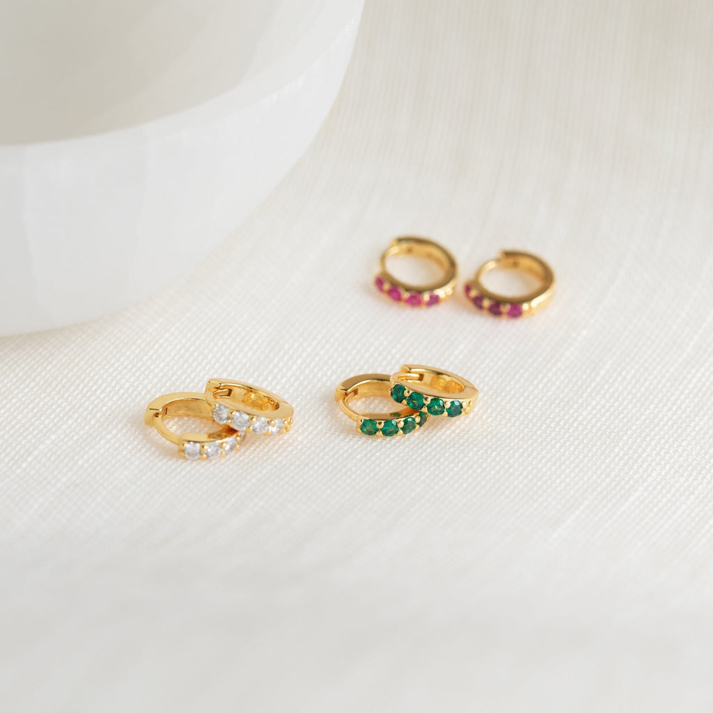 The Elise Gemstone Huggies Set features three pairs of small gold artisan-made earrings, each adorned with white, green, and pink gemstones—perfect for adding a delicate birthstone touch to any look.