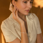 A woman in a light beige blouse wears the Lovely Bracelet (CM x Lovely Bride) and touches her ear, with a warm brown background.