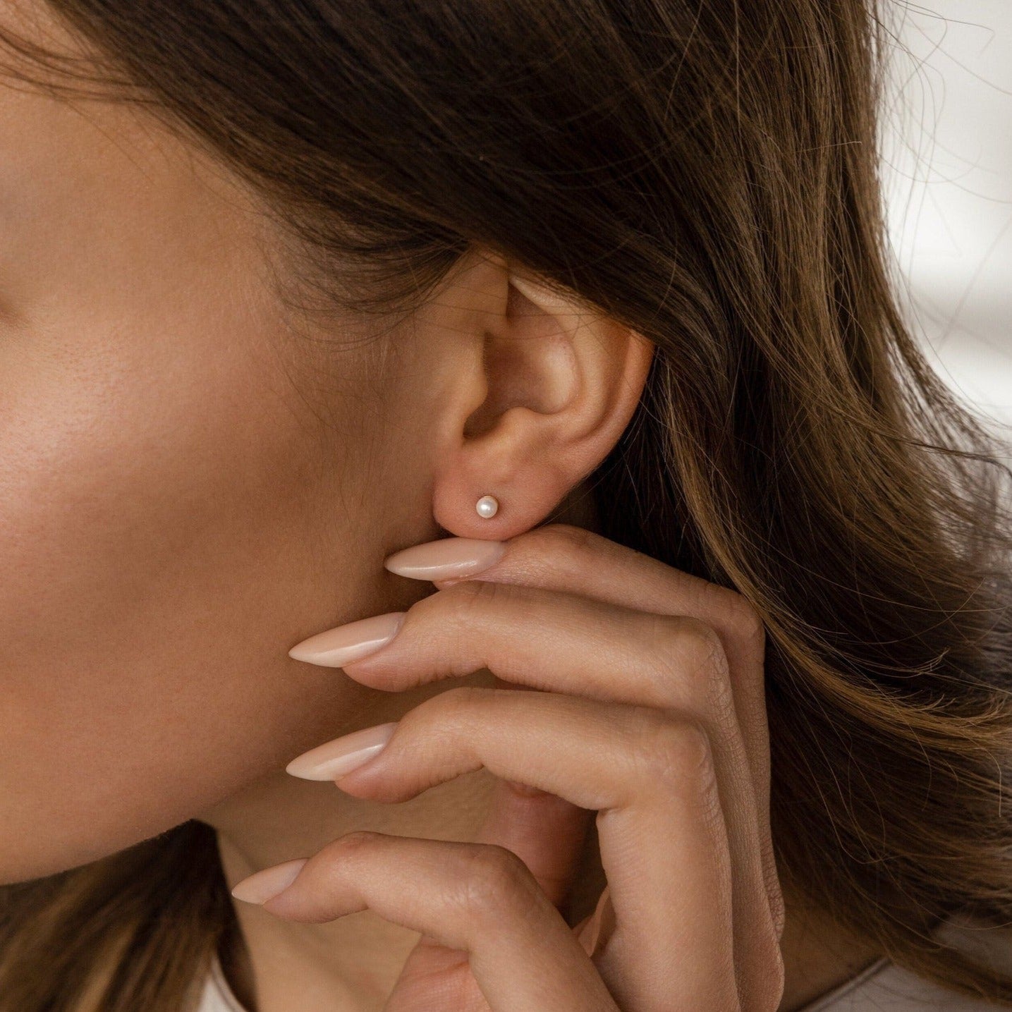 A woman with brown hair touches her ear, highlighting her nude manicured nails and wearing Dainty Pearl Studs.