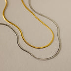 Two gold necklaces and one silver necklace from the Evergreen Necklace Set are displayed on a light beige textured surface.