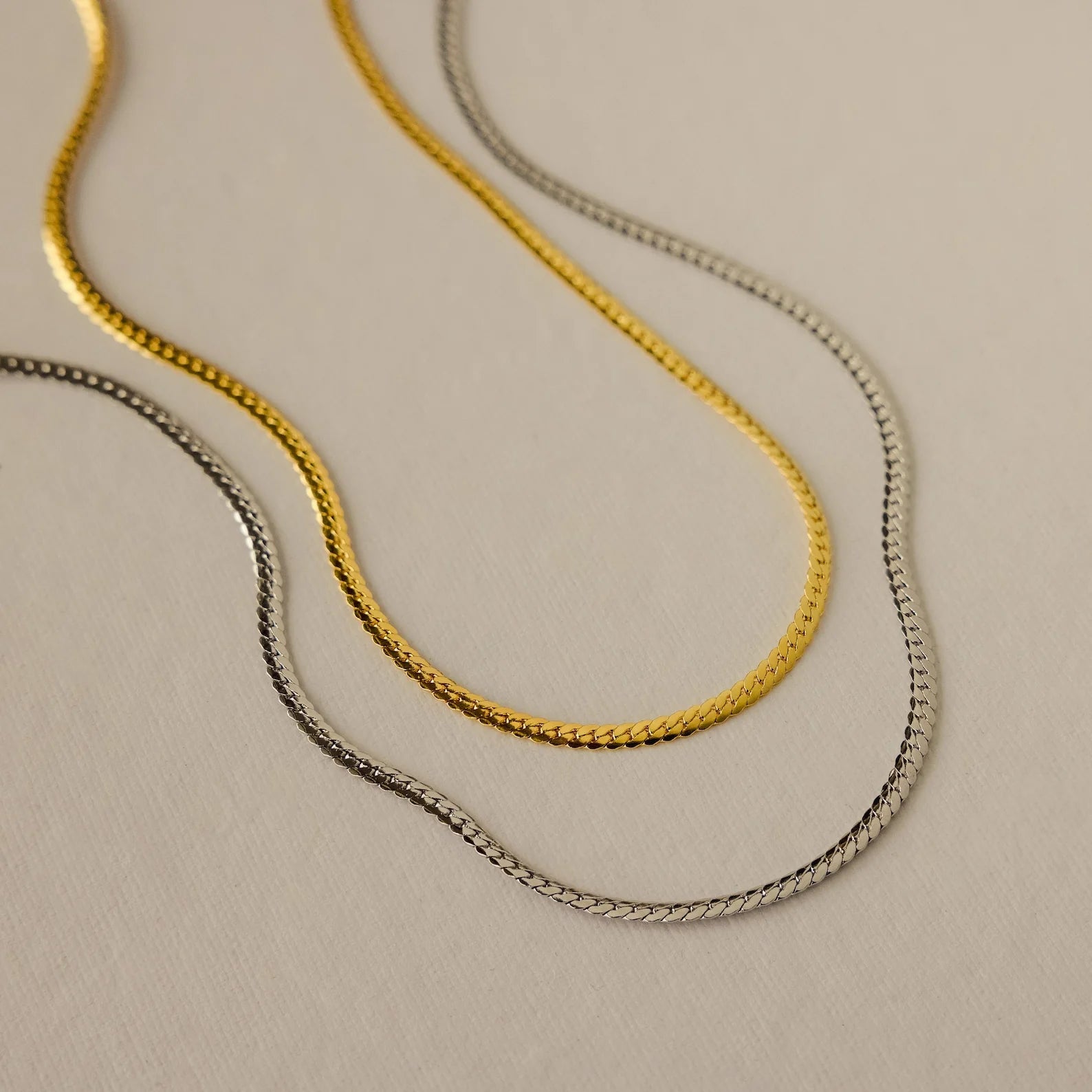 Two gold necklaces and one silver necklace from the Evergreen Necklace Set are displayed on a light beige textured surface.