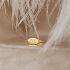 The Carrie Signet Ring, a gold minimalist piece engraved with 777, rests on a smooth surface framed by soft, white feathery textures—a unique item of personalized jewelry.