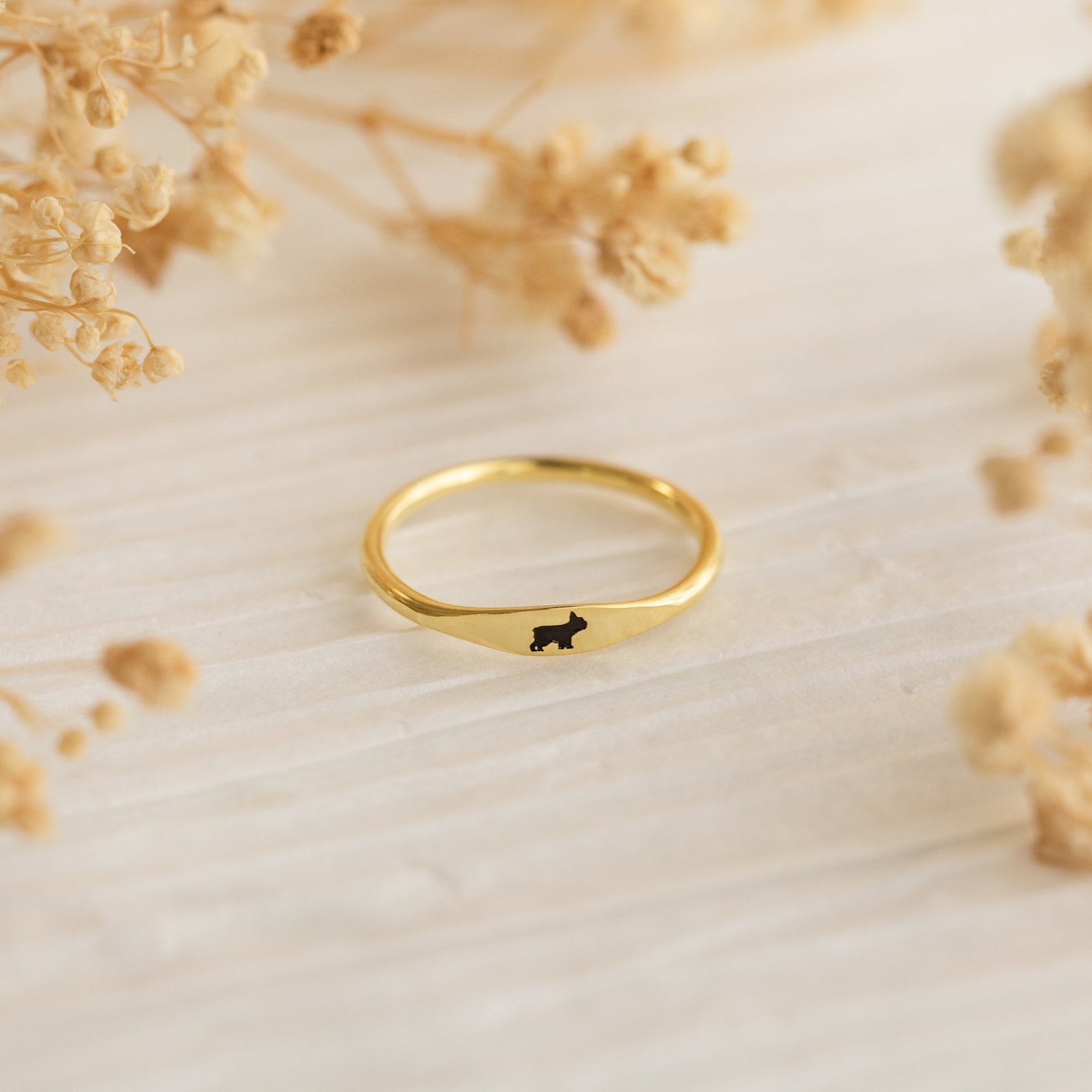 The Skinny Pet Ring—a gold stackable ring featuring a tiny black cat silhouette—rests on a light textured surface with dried flowers, making it the perfect choice for fans of dainty jewelry.