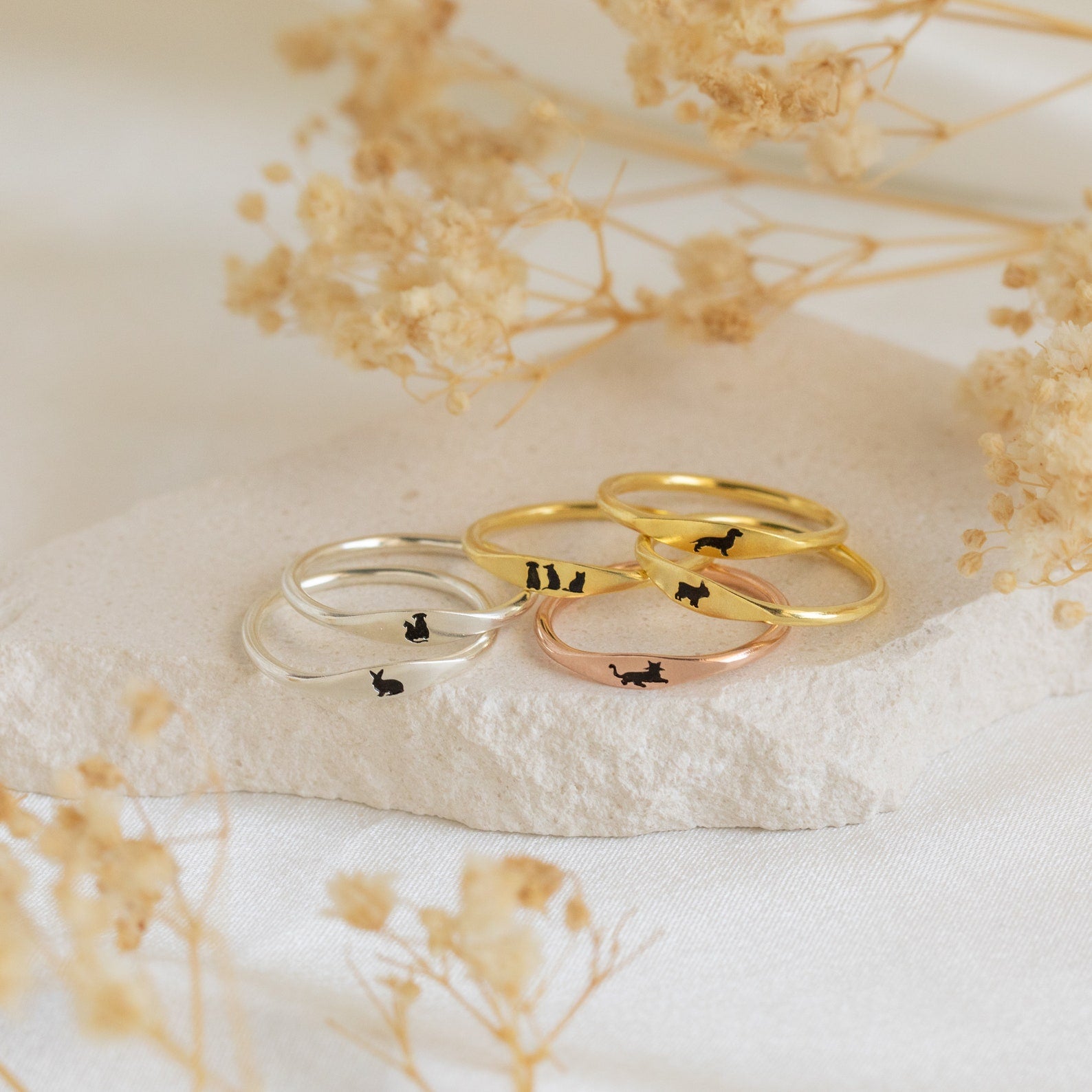 The Skinny Pet Ring set includes three gold and three silver stackable rings, each with tiny black cat silhouettes, arranged on a stone with dried flowers—ideal for fans of delicate jewelry.