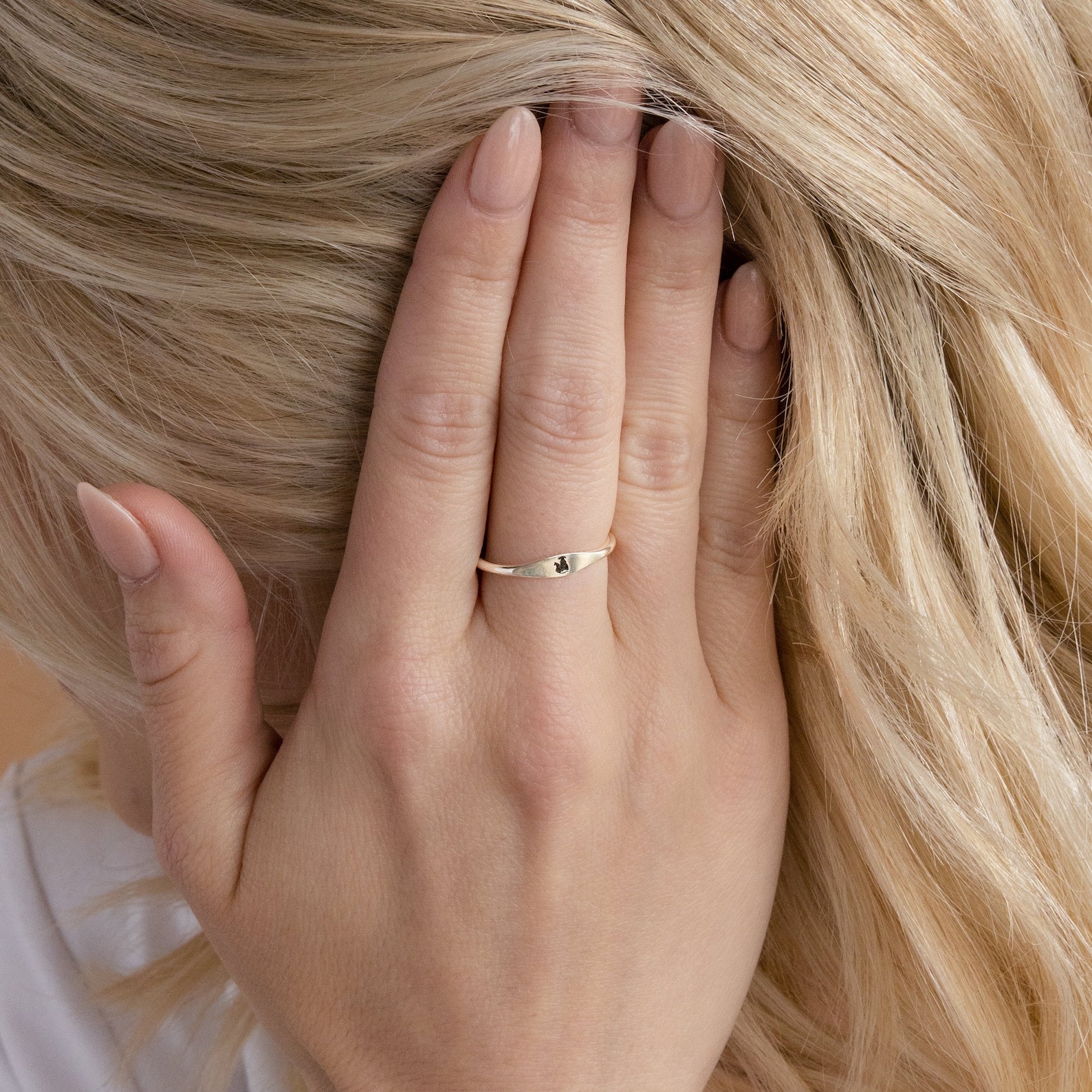 A hand with pale pink nails showcases the Skinny Pet Ring, a delicate gold stackable band featuring a small black stone, set against blonde hair.