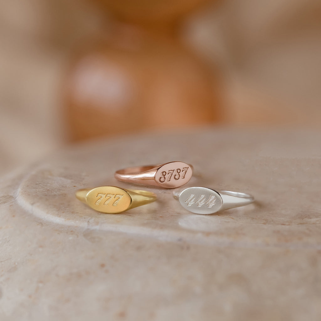 The Carrie Signet Rings—custom engraved in gold, rose gold, and silver—are displayed on a smooth beige surface, ideal for fans of personalized minimalist jewelry.
