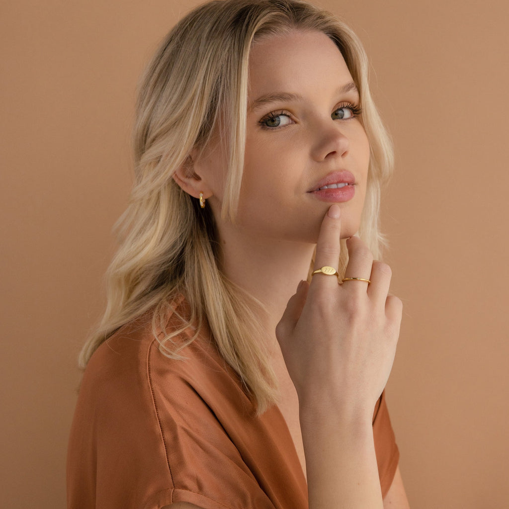 Blonde woman wearing gold jewelry, including the Carrie Signet Ring, and a brown top, poses against a beige background.