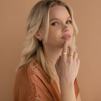 Blonde woman wearing gold jewelry, including the Carrie Signet Ring, and a brown top, poses against a beige background.