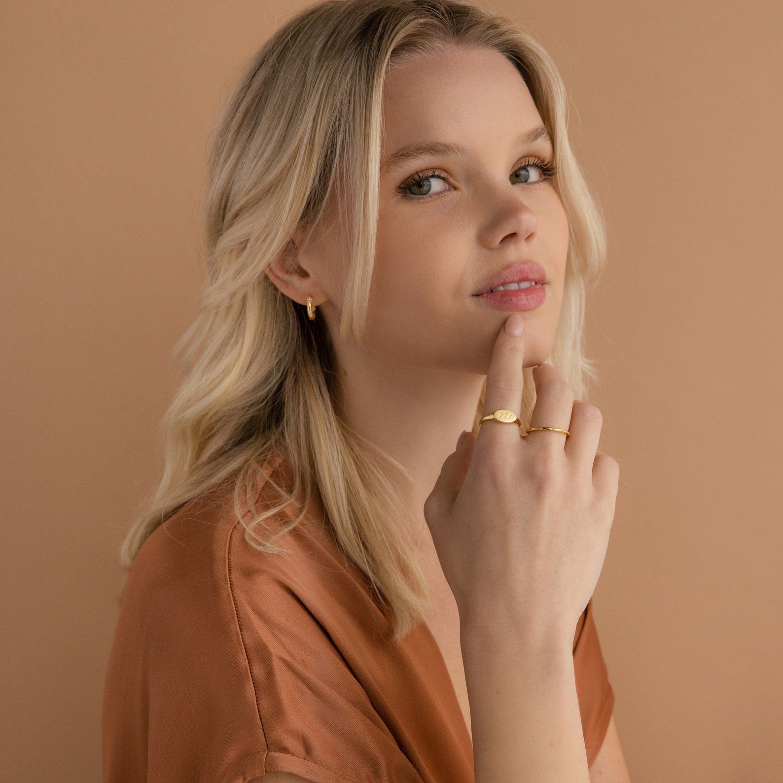 Blonde woman wearing gold jewelry, including the Carrie Signet Ring, and a brown top, poses against a beige background.