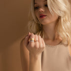 A blonde woman displays the Carrie Signet Ring alongside minimalist gold bands on her fingers, set against a beige background.