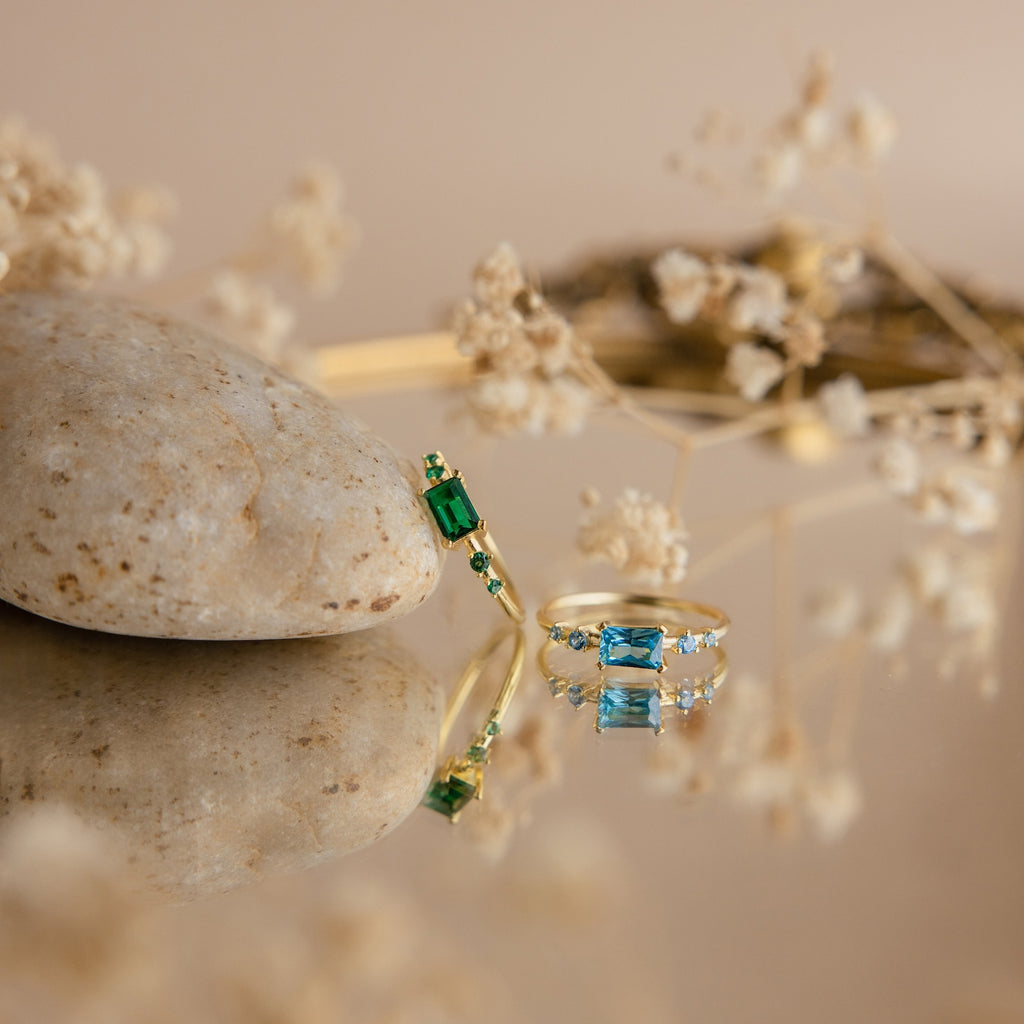 The Space Baguette Birthstone Ring—highlighting vivid green and blue stones—is displayed on a reflective surface surrounded by dried flowers and a stone.