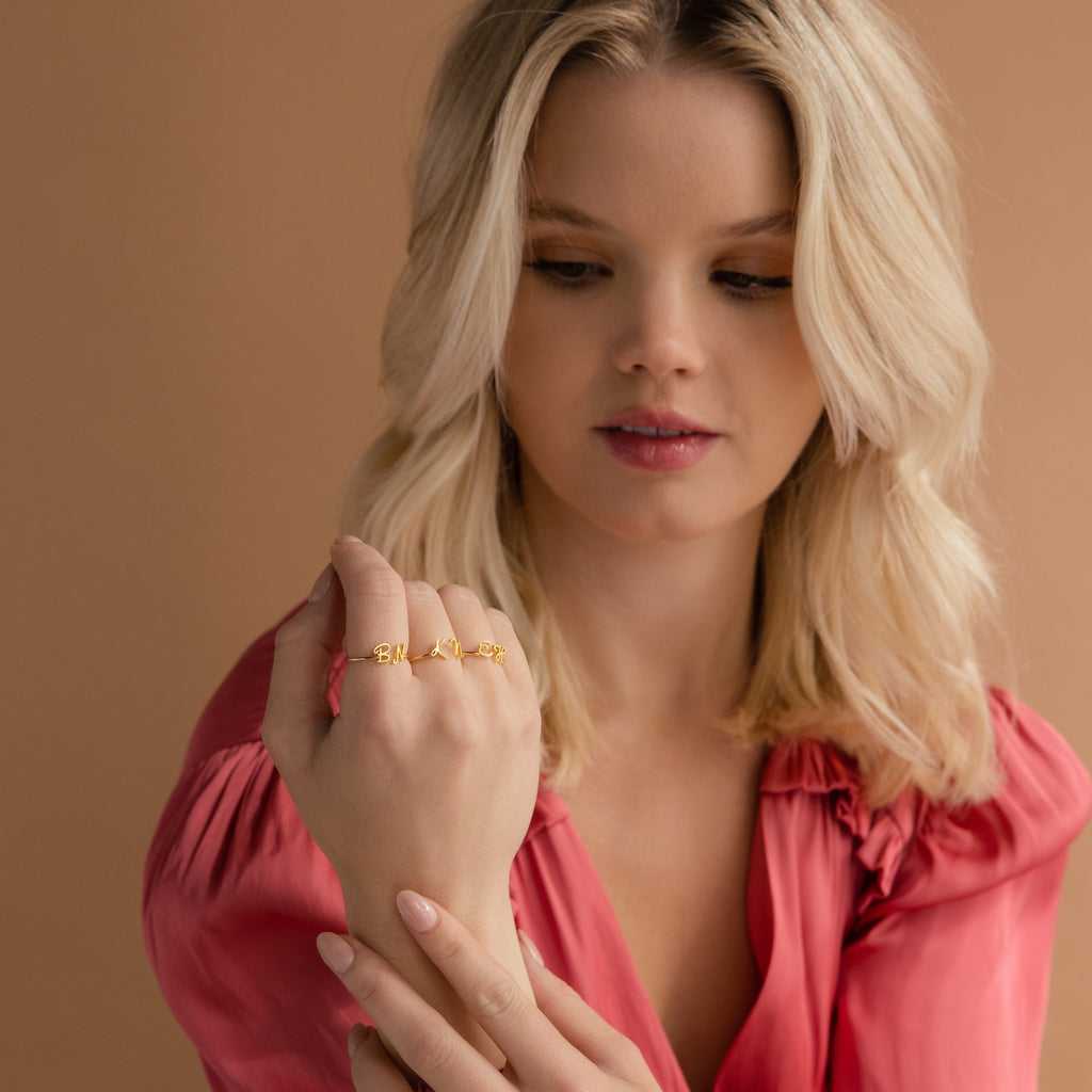 Woman in a pink blouse displays the Nella Duo Initial Ring on her fingers—an ideal personalized jewelry piece or anniversary gift—set against a neutral peach background.