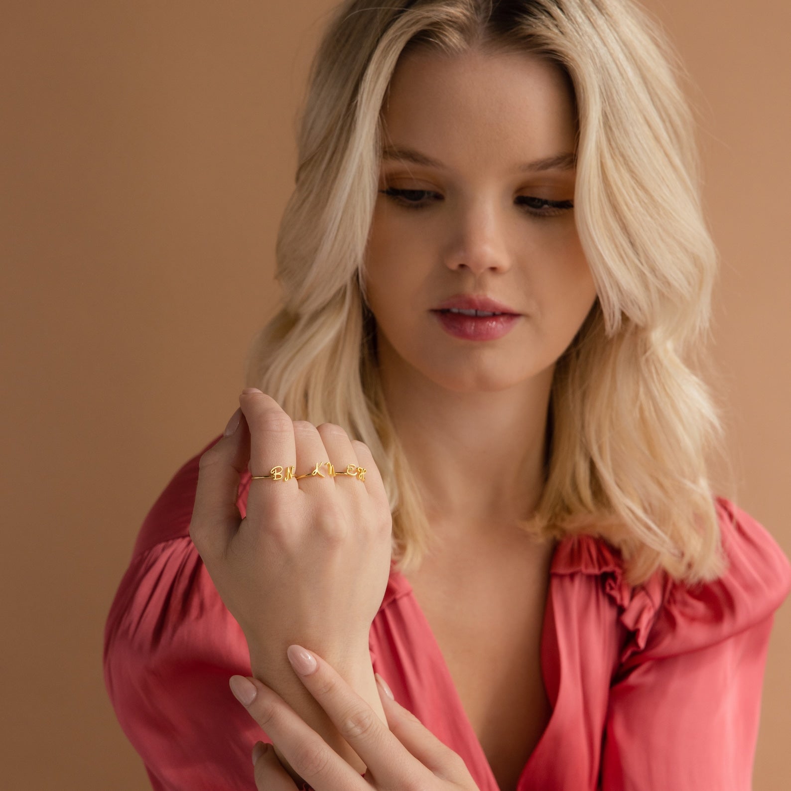Woman in a pink blouse displays the Nella Duo Initial Ring on her fingers—an ideal personalized jewelry piece or anniversary gift—set against a neutral peach background.