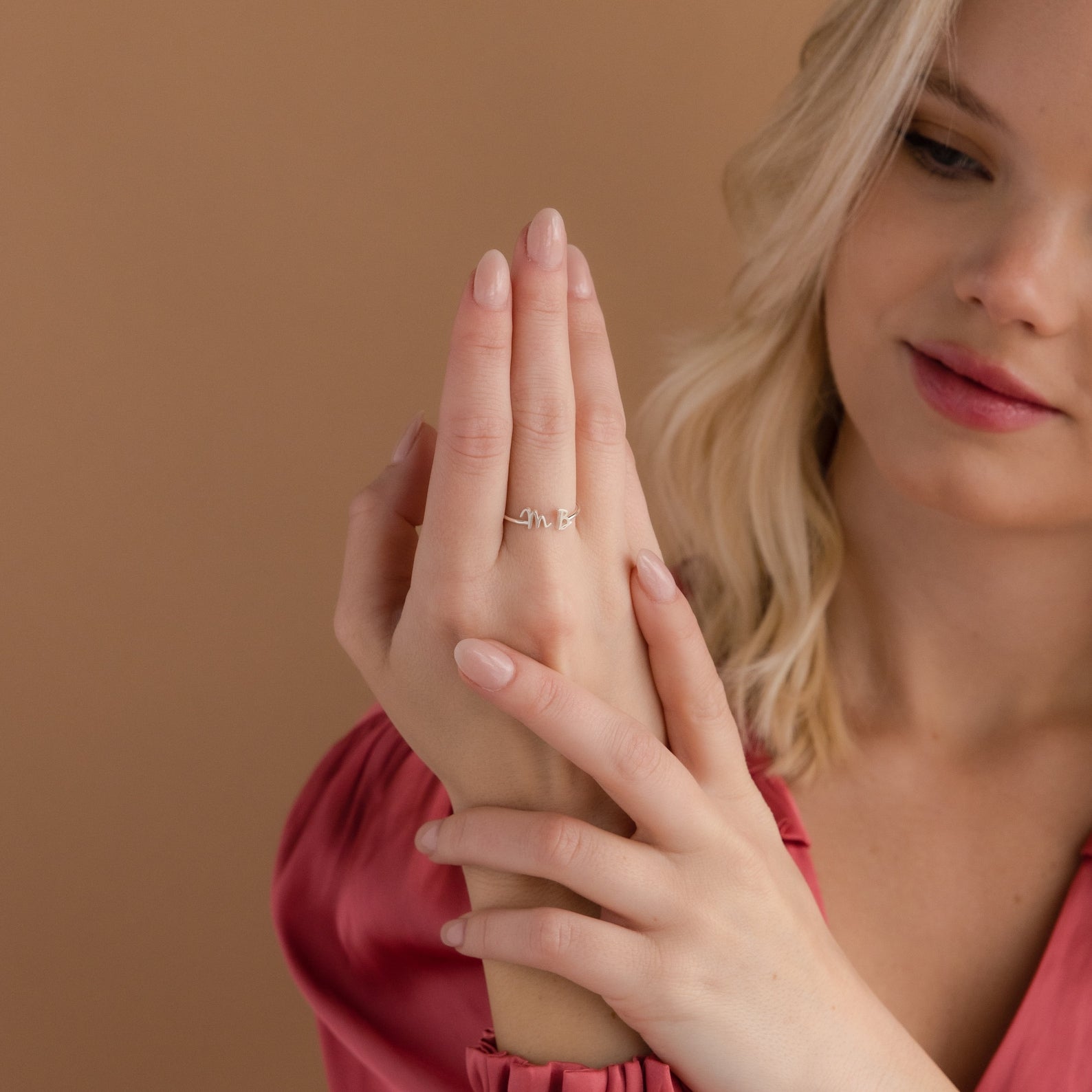 A woman with blonde hair in a pink blouse showcases the Nella Duo Initial Ring on her finger against a beige background—a lovely personalized jewelry piece ideal as an anniversary gift.