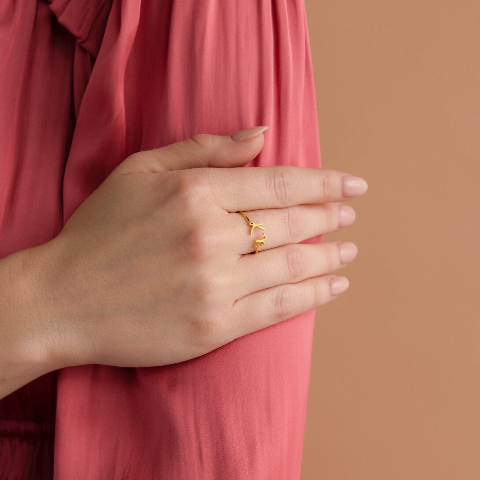 A hand wearing the Nella Duo Initial Ring rests on a pink-sleeved arm against a beige background—an elegant, personalized gift perfect for anniversaries.