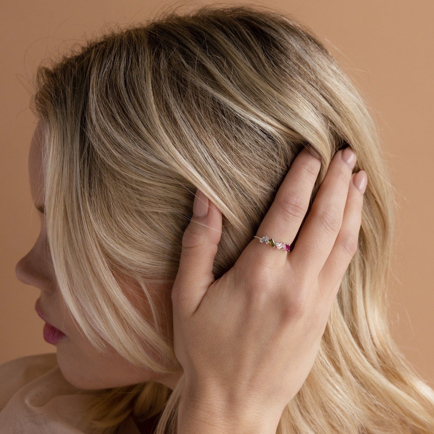 A woman with blonde hair touches her head, showcasing the Cluster Baguette Birthstone Ring in Rose Gold adorned with vibrant gemstones on her finger.