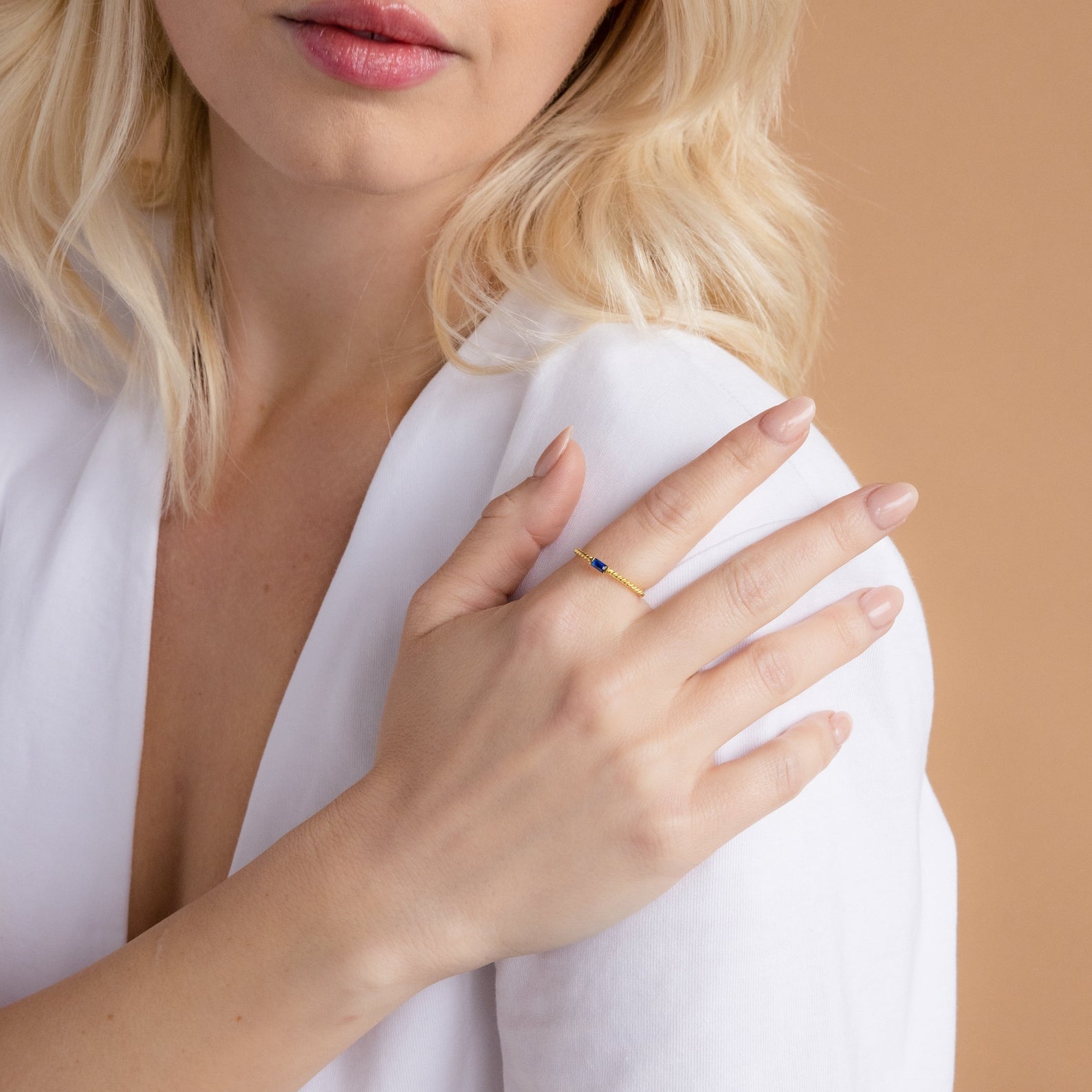 A woman with blonde hair wears a white top and the Twist Baguette Birthstone Ring featuring a small blue gem on her finger.