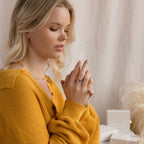 A woman in a mustard sweater admires the Mothers Baguette Birthstone Ring on her hand, with jewelry boxes on the table beside her.