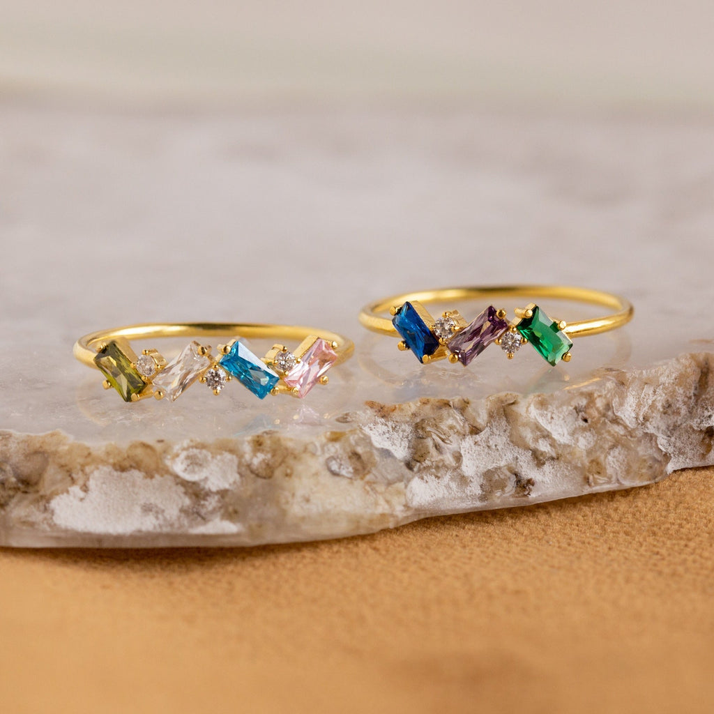 The Cluster Baguette Birthstone Ring features colorful rectangular and round gemstones in gold, ideal for a personalized statement or birthstone ring, beautifully displayed on marble and stone surfaces.