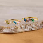 The Cluster Baguette Birthstone Ring features colorful rectangular and round gemstones in gold, ideal for a personalized statement or birthstone ring, beautifully displayed on marble and stone surfaces.