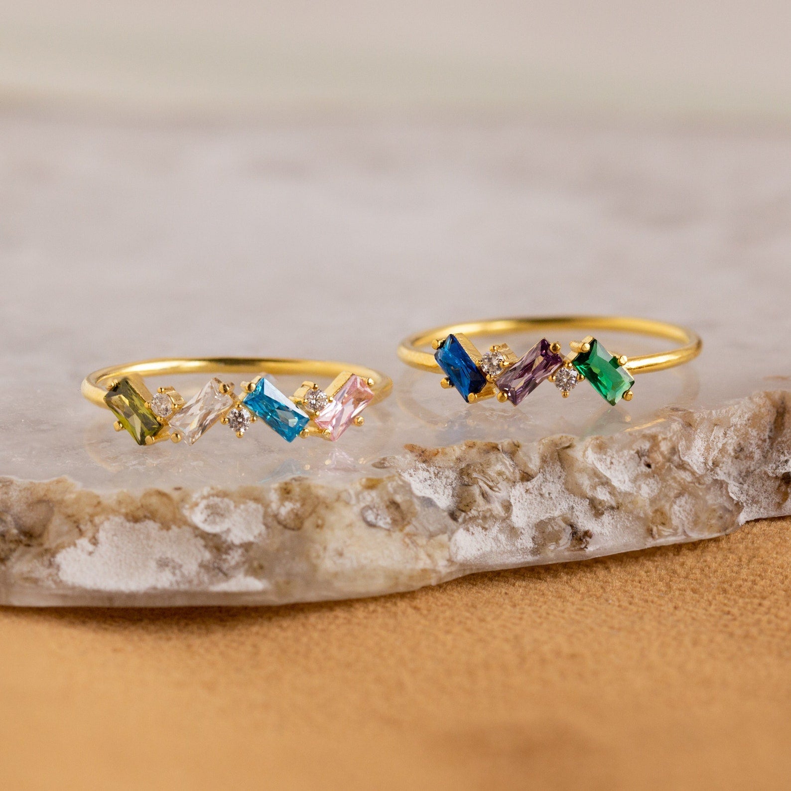 The Cluster Baguette Birthstone Ring features colorful rectangular and round gemstones in gold, ideal for a personalized statement or birthstone ring, beautifully displayed on marble and stone surfaces.