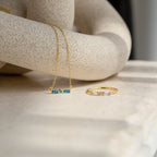 A gold chain and ring rest on a marble surface, complemented by an Initial Morse Birthstone Necklace for a subtle touch of personalized jewelry.