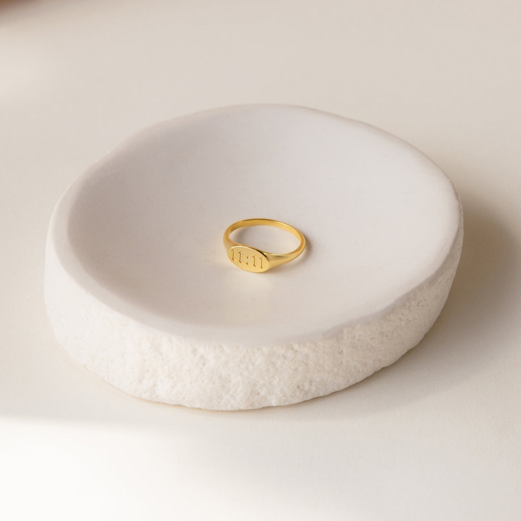 The Old English Signet Ring, engraved with 1111, sits in a round white ceramic dish on a light background—a subtle and personalized piece of jewelry.