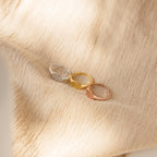 The Old English Signet Ring, available in silver, gold, and rose gold finishes, is displayed on soft beige textured fabric—a perfect minimalist piece for those who appreciate personalized jewelry with subtle elegance.