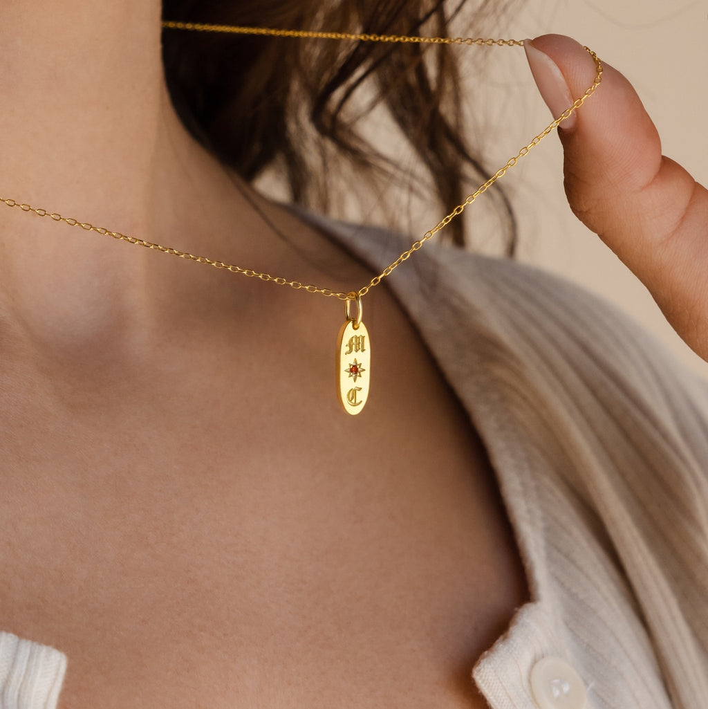 A woman wears a beige top and holds the Old English Oval Tag Necklace—a gold pendant personalized with initials and a starburst engraving.