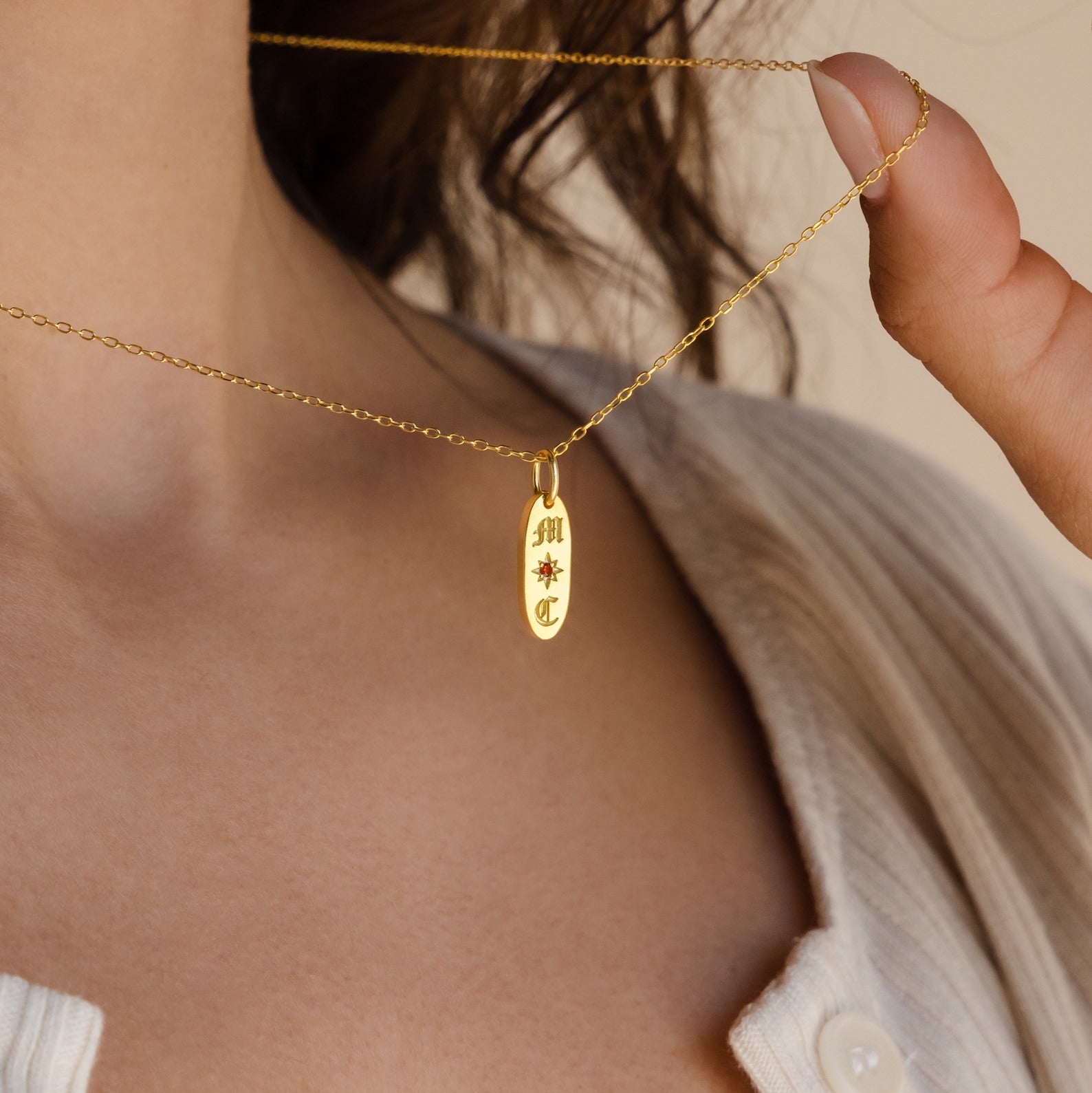 A woman wears a beige top and holds the Old English Oval Tag Necklace—a gold pendant personalized with initials and a starburst engraving.