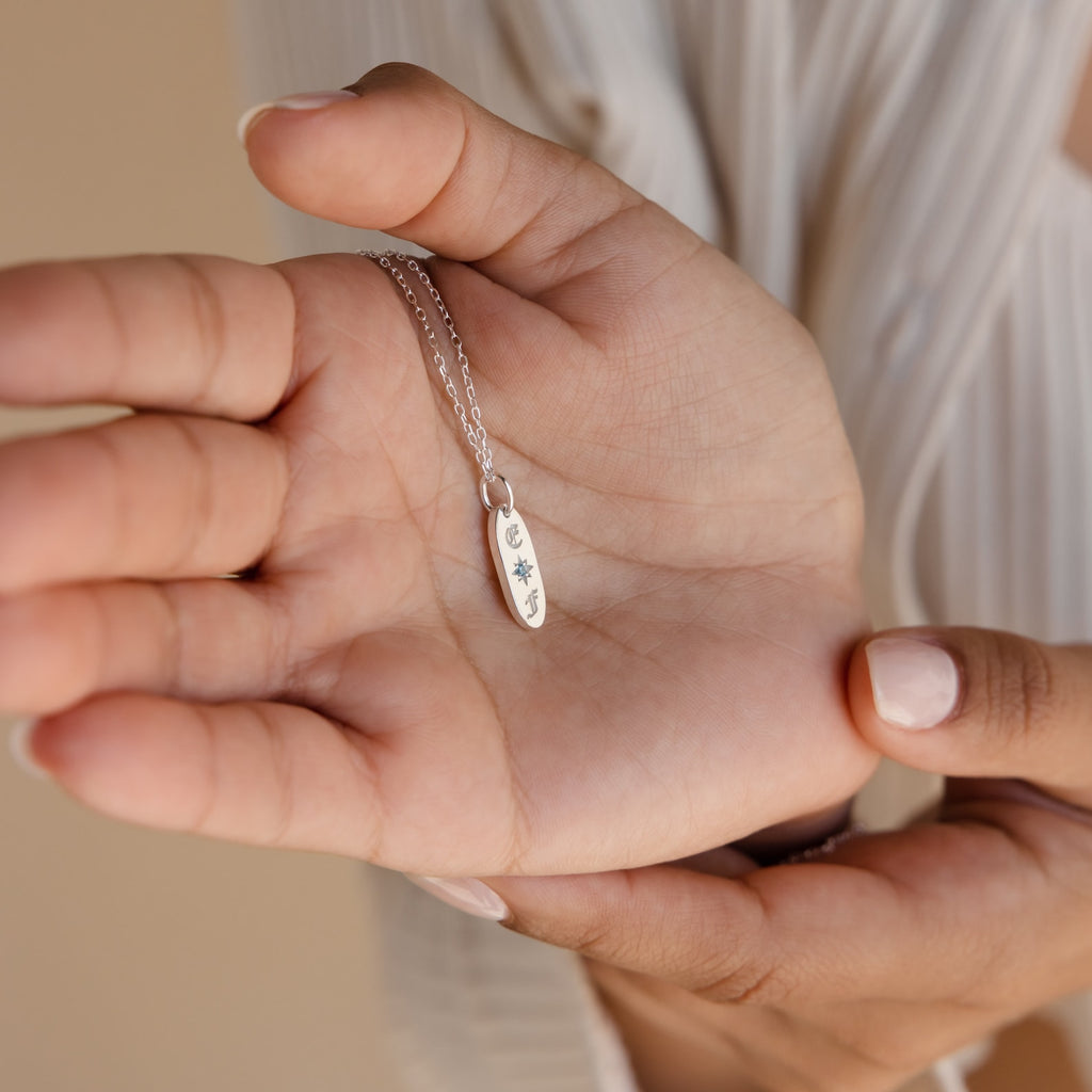 A person holds the Old English Oval Tag Necklace, a silver pendant engraved with crescent moon, star, and sun symbols plus delicate personalized initials.
